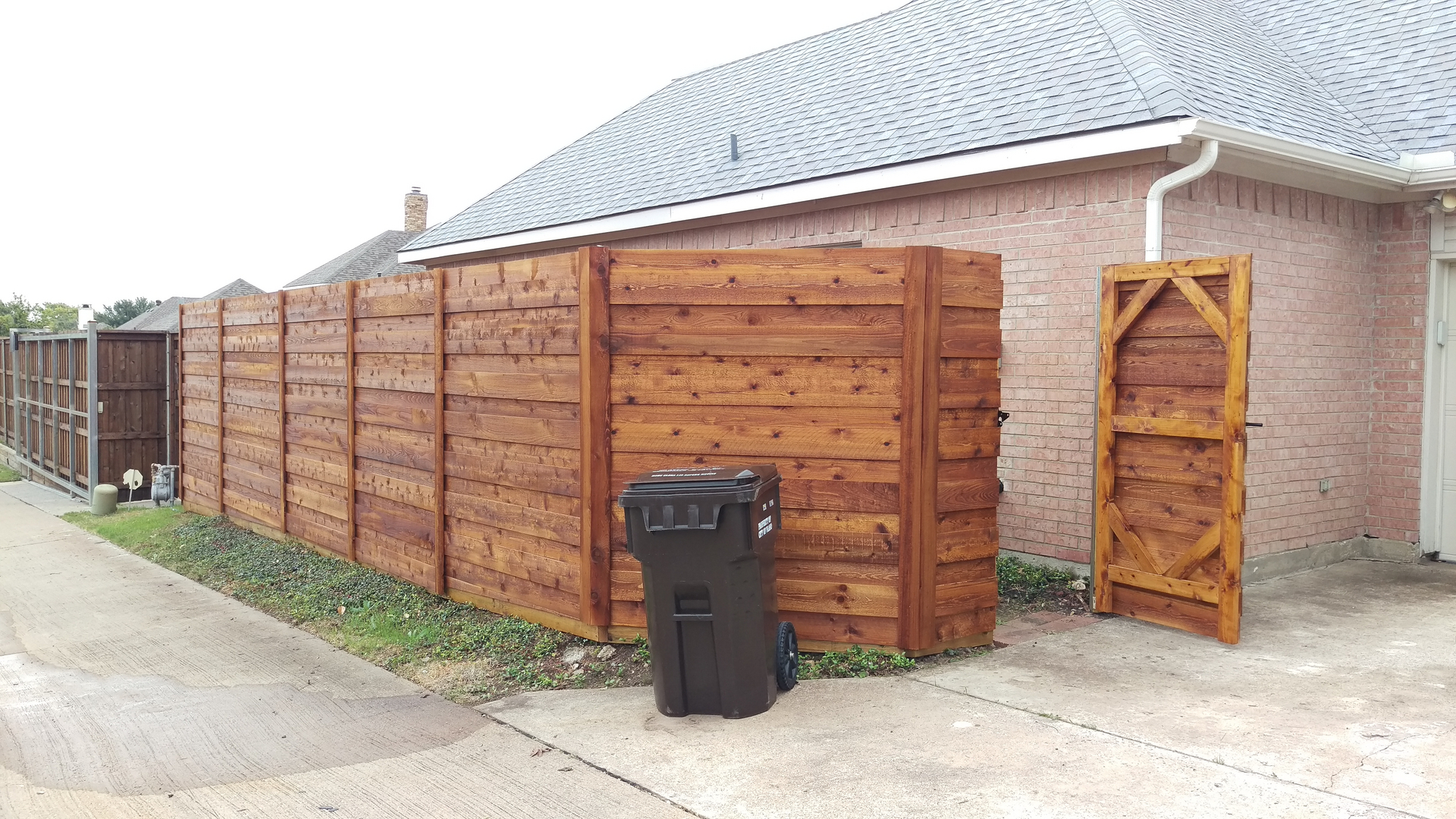 A black trash can sitting in front of a wooden fence.