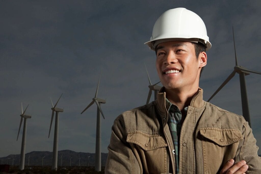 A man in a white hard hat standing next to wind mills.